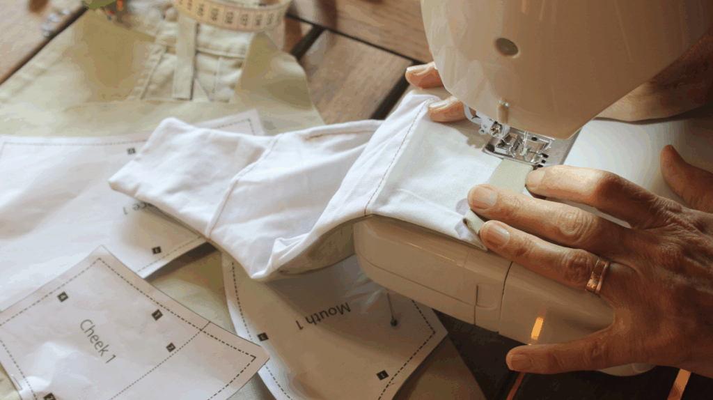 Garment sample being sewn during early development stage Close-up of hands sewing a garment piece on a machine, with fabric and paper pattern templates on a wooden table โ suggesting early-stage product development or prototyping