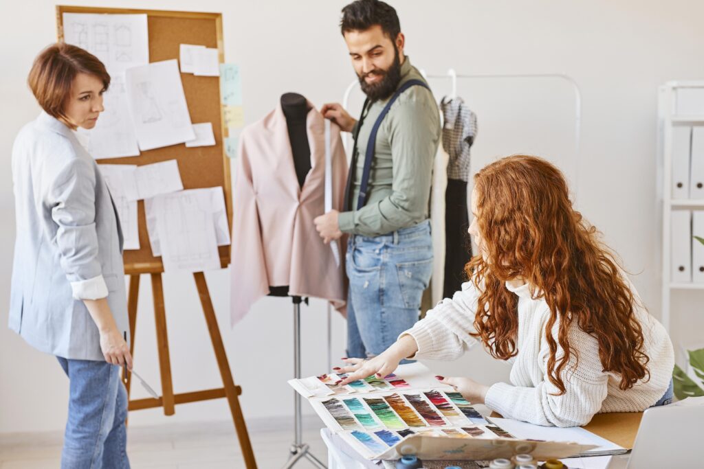 Three fashion designers reviewing a dress form with color palettes in a studio, illustrating garment sampling and approval steps before bulk production.