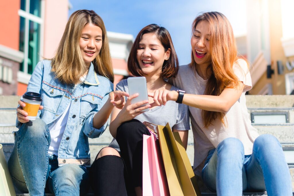 Three young women looking at a smartphone while shopping, illustrating Gen Z and Gen Alpha fashion spending and how youth influence trend visibility and buying decisions.